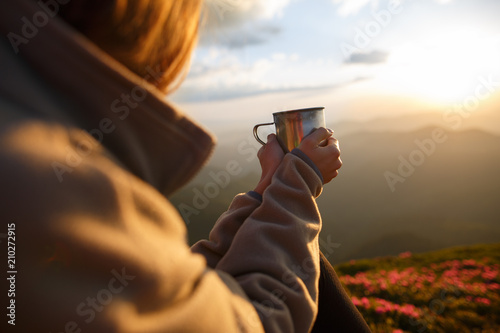 Closeup photo of cup with tea in traveler's hand over out of focus mountains view. A young tourist woman drinks a hot drink from a cup and enjoys the scenery in the mountains. Trekking concept
