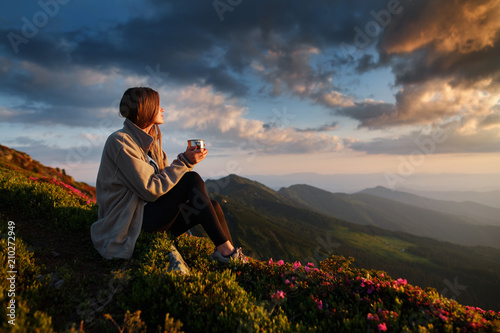 woman traveler drinks coffee with a view of the mountain landscape. A young tourist woman drinks a hot drink from a cup and enjoys the scenery in the mountains. Trekking concept