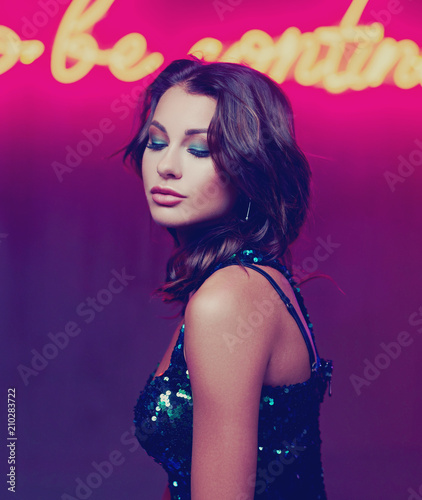 Fashion portrait of young beautiful woman with middle length dark hair wearing green sequins dress posing against red wall with neon letters in night bar © Dmitry Tsvetkov