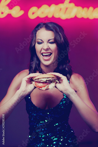Sexy gorgeous woman in night dress with sequins eating hamburger in night club. Girl standing and posing against red wall with neon letters