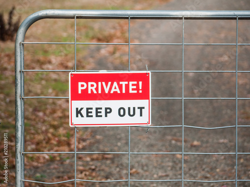A sign says ' Private! Keep out ' on a iron gate with dirt road driveway at the background.