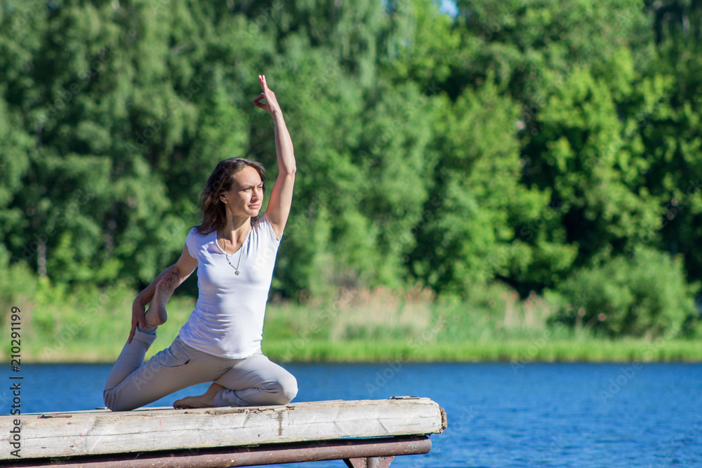 Young caucasian woman doing yoga asanas outdoors on a wooden pier by the river or lake, sunny summer morning, wearing no makleup and sports clothes