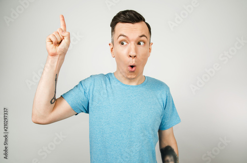 Young thinking guy dressed in a blue t-shirt shows finger up with wide eyes and opened mouth on a light background. © vika33