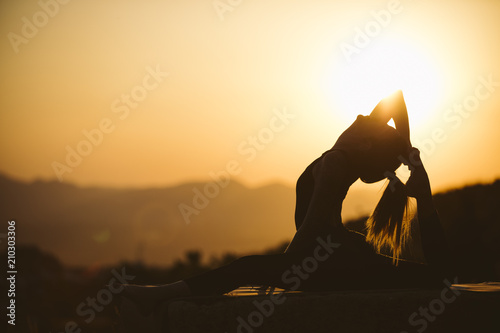 Fototapeta Naklejka Na Ścianę i Meble -  Young woman is practicing yoga on the mountain at sunset. Silhouette of young woman practicing yoga outdoor.