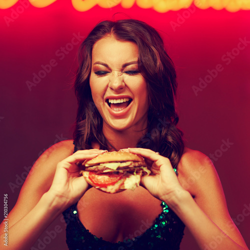 Sexy gorgeous woman in night dress with sequins eating hamburger in night club. Girl standing and posing against red wall with neon letters