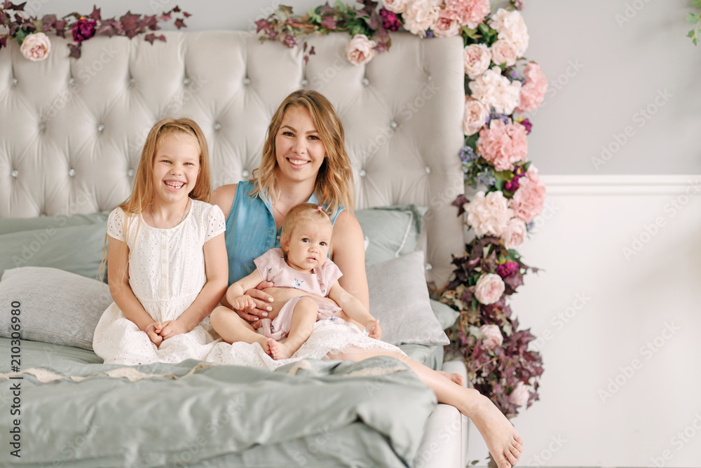 A happy mother with two beautiful daughters is sitting on the bed. They laugh and hug each other A happy mother with two beautiful daughters is sitting on the bed. They laugh and hug each other
