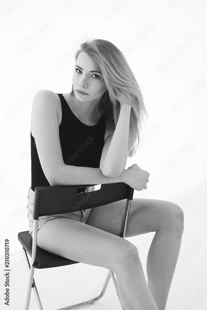 Young beautiful sexy woman in blue jeans shorts and black shirt posing and sitting on simple black chait in white studio. Fashion model with long straight hair. Young beautiful sexy woman in blue jeans shorts and black shirt posing and sitting on simple black chait in white studio. Fashion model with long straight hair.