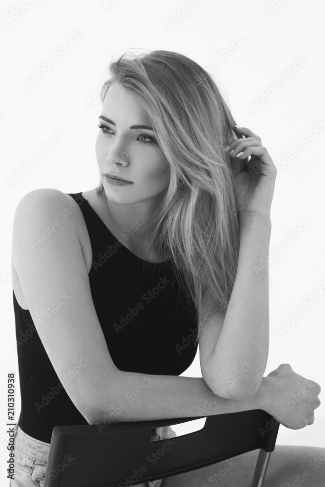 Young beautiful sexy woman in blue jeans shorts and black shirt posing and sitting on simple black chait in white studio. Fashion model with long straight hair. Young beautiful sexy woman in blue jeans shorts and black shirt posing and sitting on simple black chait in white studio. Fashion model with long straight hair.