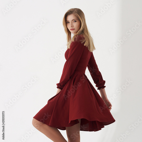 Young blonde woman in cherry red dress standing and posing on white background. Female with long straight hair.