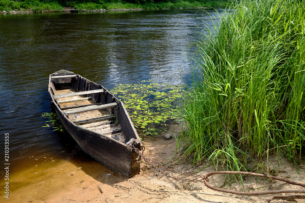 Old wooden rowing boat tied with chain and padlock at green summer bank on Neman river in Grodno Belarus Old wooden rowing boat tied with chain and padlock at green summer bank on Neman river in Grodno Belarus