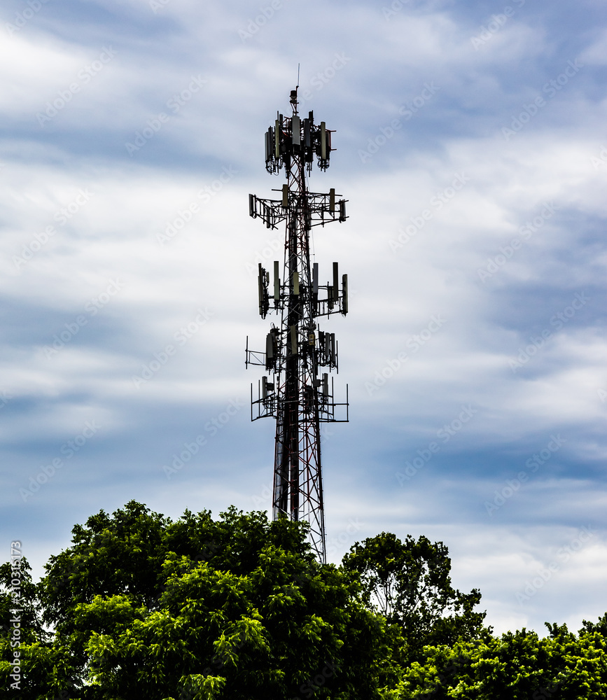A cell phone tower piercing the greenery below, set against a cloudy blue sky.