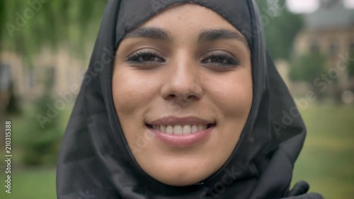 Face of young beautiful muslim girl in hijab is standing and smiling in daytime, in bad weather, watching at camera, religiuos concept, blurred background photo