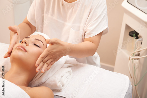 Young beauty specialist touching cheeks of client to calm skin down after skincare procedure