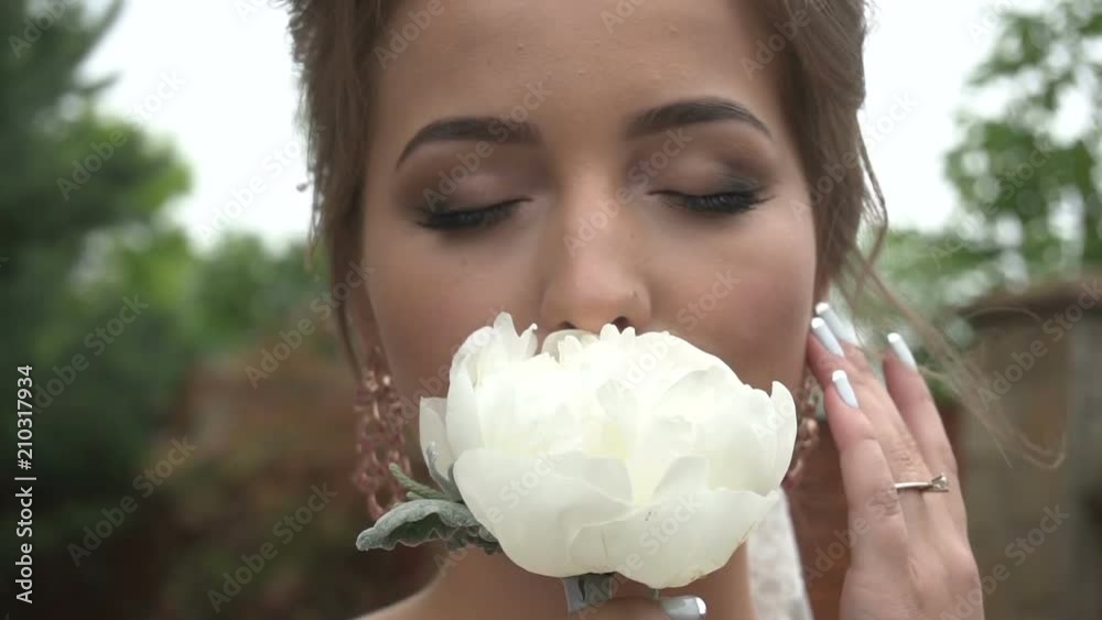 Close-up portrait of the charming bride with natural make-up smelling the white peony with closed eyes.