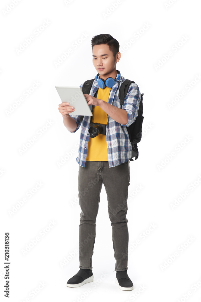 Full length portrait of concentrated Asian man with backpack standing against white background and studying map on digital tablet