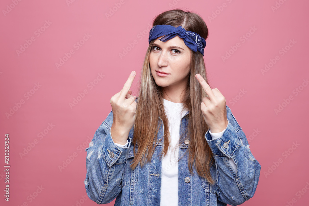 Irritated hippy female being fed up with relations with lover, keeps hands in fuck you sign, wears fashionable denim clothes in hippie style, isolated over pink background. Annoyance and subculture. Irritated hippy female being fed up with relations with lover, keeps hands in fuck you sign, wears fashionable denim clothes in hippie style, isolated over pink background. Annoyance and subculture.