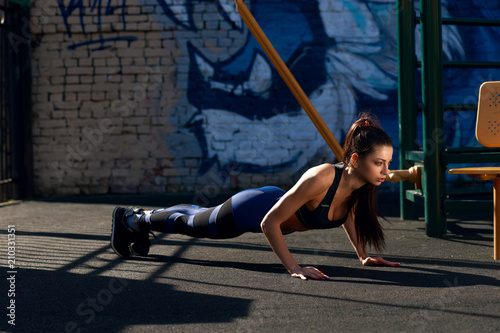 Fitness model exercising. Pretty tanned woman in top and leggings doing push ups at outdoor workout space. Sport and health concept.