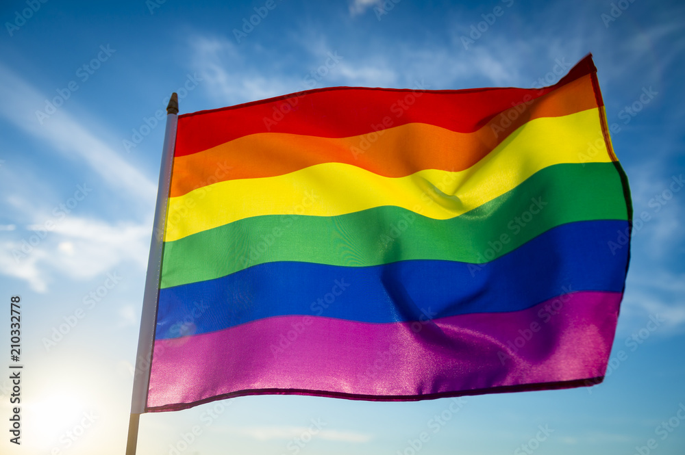 Obraz premium Backlit gay pride rainbow flag flutters in the breeze with lens flare.