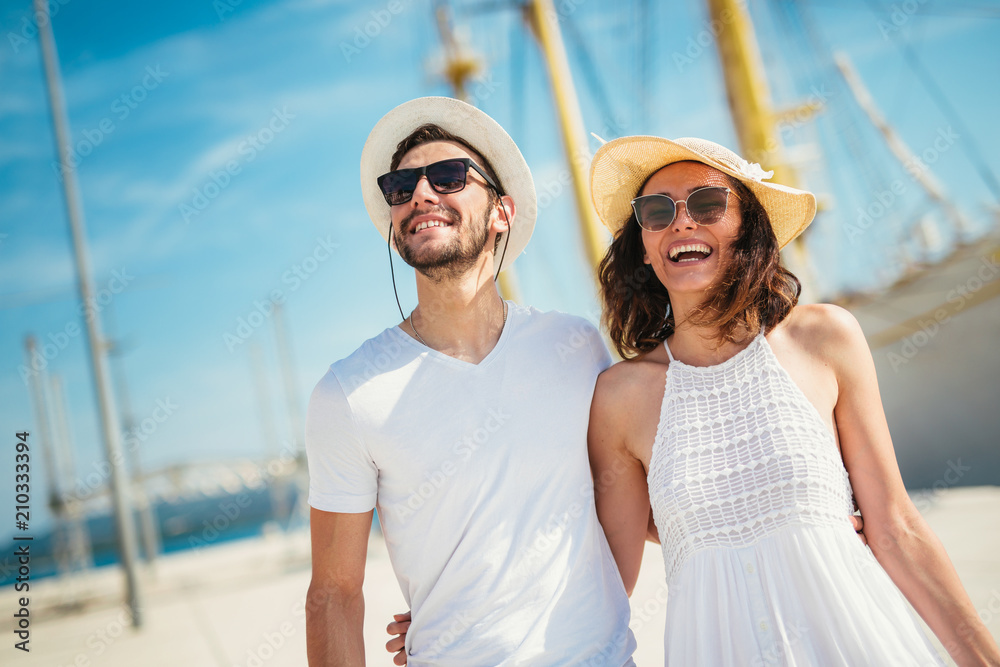 Happy young couple walking by the harbor of a touristic sea resort with sailboats on background Happy young couple walking by the harbor of a touristic sea resort with sailboats on background