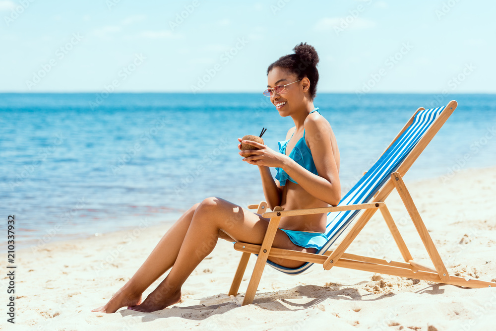 happy african american woman in bikini and sunglasses drinking cocktail in coconut shell while sitting on deck chair on sandy beach