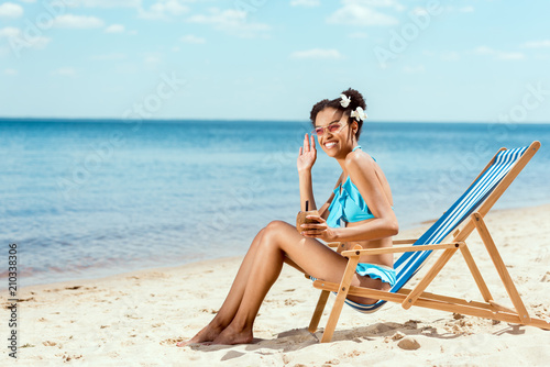 happy african american woman in bikini with cocktail in coconut shell waving by hand while sitting on deck chair on sandy beach