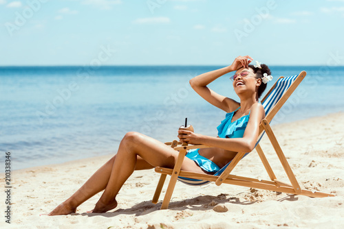Foto young african american woman in bikini wiping forehead and holding cocktail in c