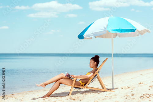 side view of african american woman reading magazine and relaxing on deck chair under beach umbrella in front sea