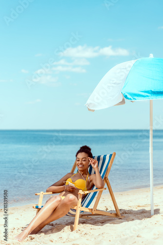 smiling african american woman relaxing on deck chair and listening music in earphones with smartphone under beach umbrella in front of sea