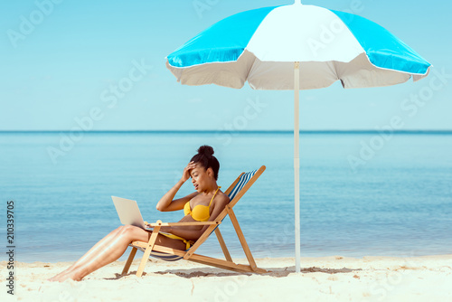 side view of smiling african american female freelancer sitting on deck chair and using laptop under beach umbrella in front of sea