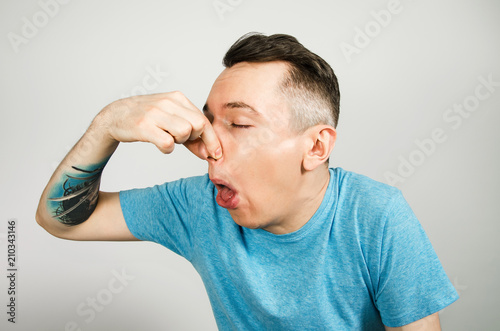 Young guy closing his nose from a bad smell, dressed in a blue t-shirt on a light background. photo