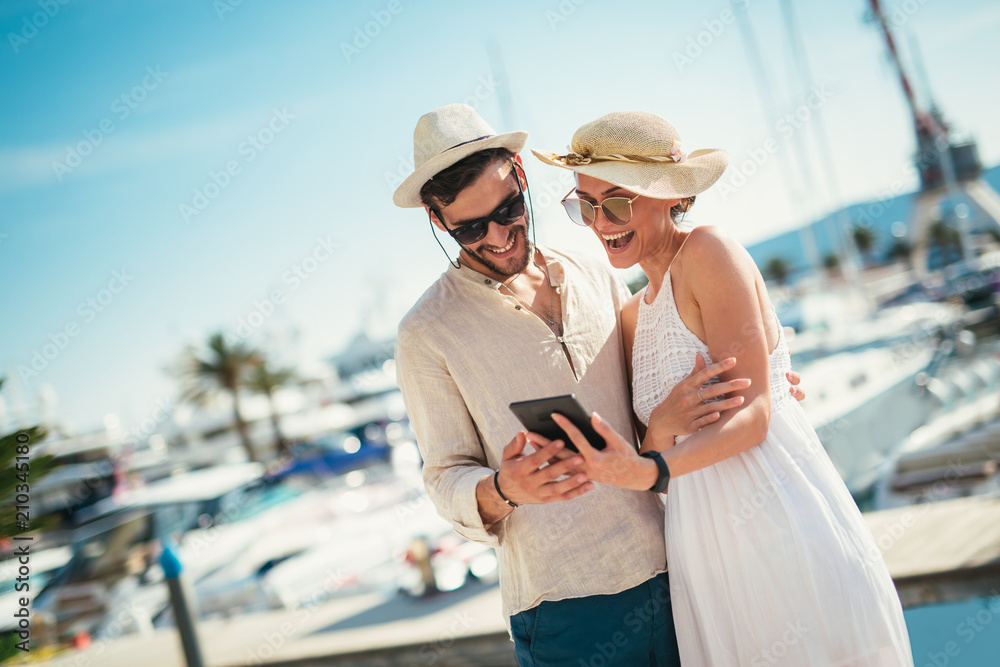 Happy young couple walking by the harbor using digital tablet of a touristic sea resort with sailboats on background