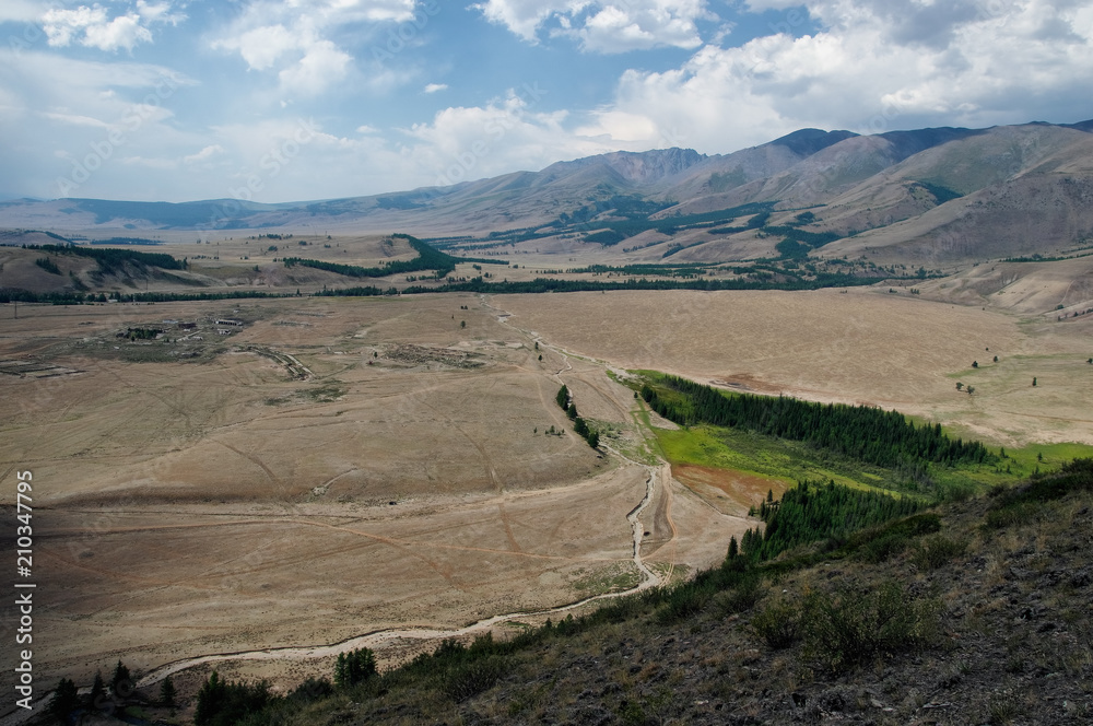 Road through a dry desert steppe on a highland mountain plateau with yellow grass trees with ranges of  hills rocks on a horizon skyline Kurai Altai Mountains Siberia Russia