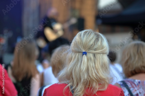 Blondynka, kobieta, stoi tyłem, przed nią, rozmyty, tłum ludzi, na scenie występuje gitarzysta, koncert, wydarzenie publiczne