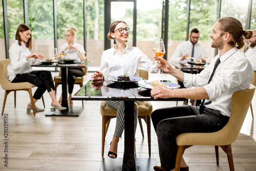 Beautiful couple clinking with wine glasses during a business lunch with delicious meals at the modern restaurant with business people on the background