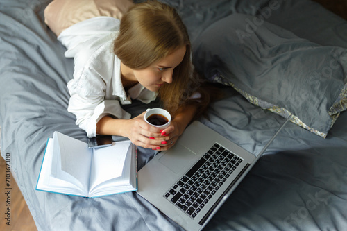Young girl in a white shirt, with a cup of coffee lying on the bed and working on the computer. Woman wake up and work with laptop and smartphone start project on bed in bedroom, early morning