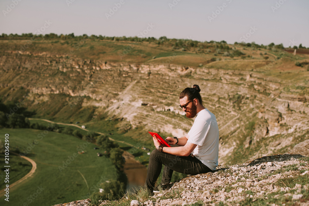 Bearded stylish young man with long hair looking on and holding a  tablet at sunset on a field. Technology, travel and modern life concept.