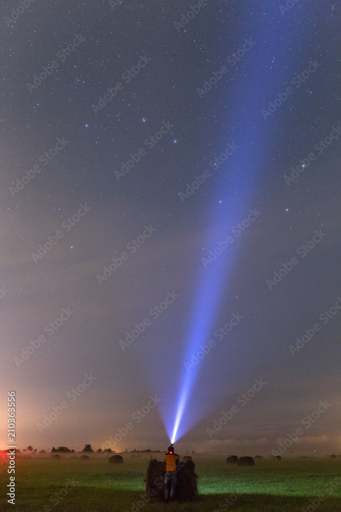 A man staying near the haystack in the green field at night.  Light ray pointing to stars