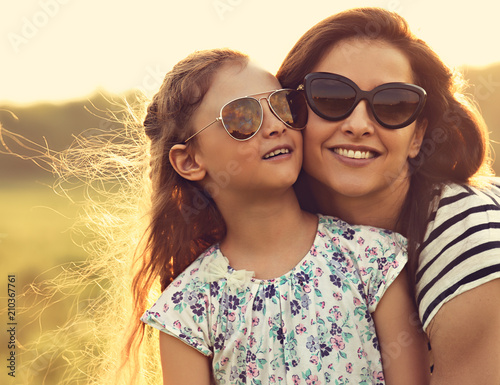 Happy fashion kid girl embracing her mother in trendy sunglasses and looking on nature background. Closeup toned portrait
