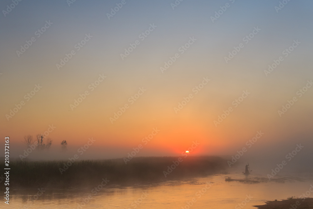 Two fishermen swimming in a boat on a foggy river under the rising sun at dawn