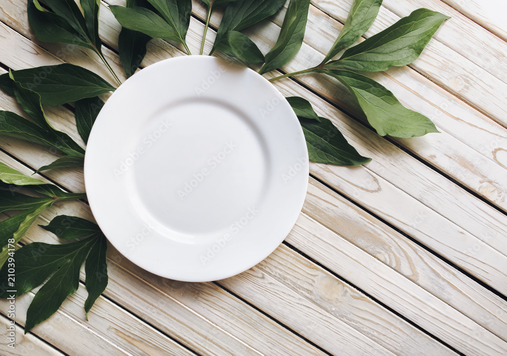 Empty white plate on light gray wooden background with green leaves of peony. Top view, copy space, close up. Empty white plate on light gray wooden background with green leaves of peony. Top view, copy space, close up.