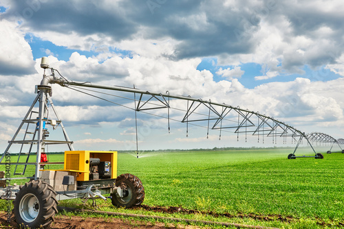 irrigation machine watering agricultural field with young sprouts, green plants on black soil and beautiful sky