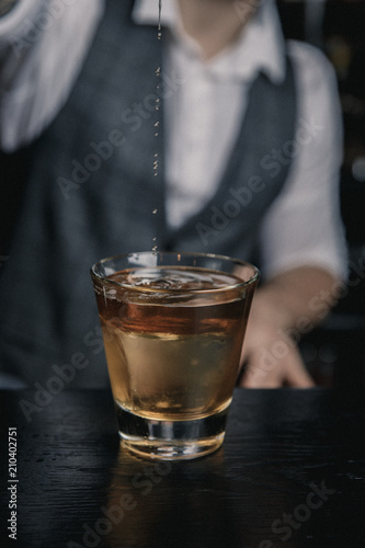 Barman in a white shirt and gray waistcoat, pouring a cocktail into the rocks glass, with a big ice cube in it.