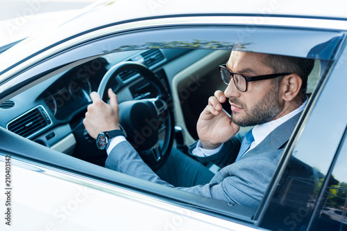 side view of businessman in eyeglasses talking on smartphone while driving car