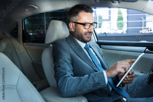 side view of smiling businessman in eyeglasses using tablet in car