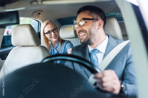 selective focus of smiling businessman in eyeglasses driving car with colleague sitting behind on passengers seat