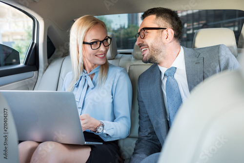 portrait of smiling business people with laptop on back seats in car