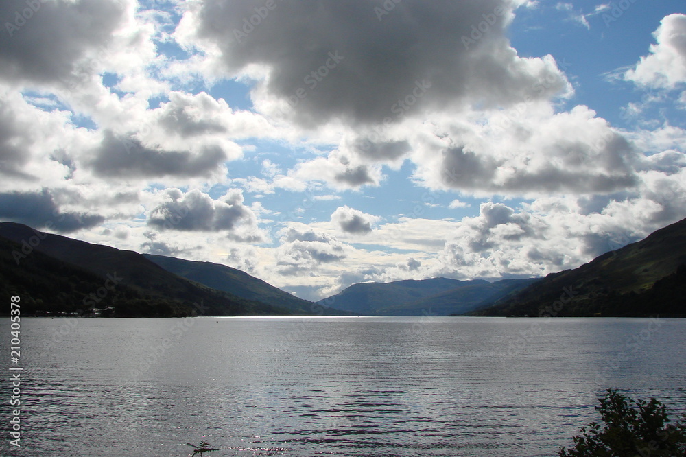 Panorama of cloudy blue northern sky at sunset, reflected on the mirror surface of Scottish lake, surrounded by hills.