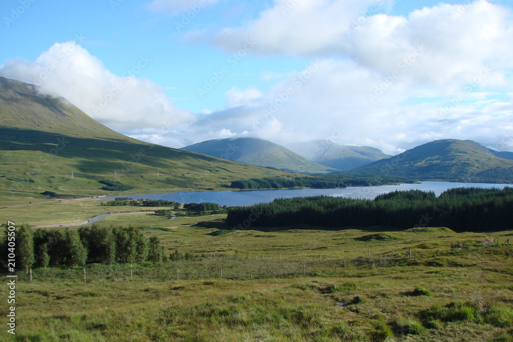 The landscape of Scottish forests under the sun's rays of the morning sun near the blue lake and at the foot of low mountains.