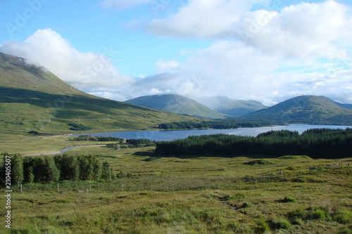 The landscape of Scottish forests under the sun's rays of the morning sun near the blue lake and at the foot of low mountains.