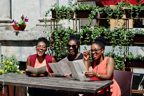 Three stylish african american womans posed at sunny summer day outdoor, sitting on table of restaurant with menu at hands.